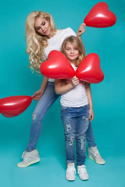 Beautiful blonde mother and daughter stand on a blue background and holding red Valentine's balloons — Stock Photo, Image