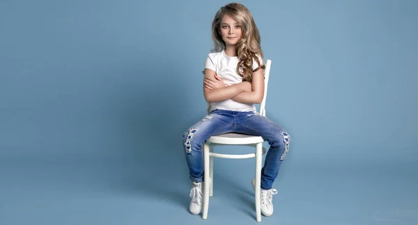 Krásné, úžasné málo blonďaté dívky v džíny sedí na židli na modrém pozadí — Stock fotografie