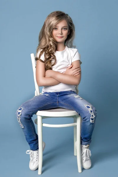 Vackra fantastisk liten blond flicka i denim jeans sitter på en stol på en blå bakgrund — Stockfoto