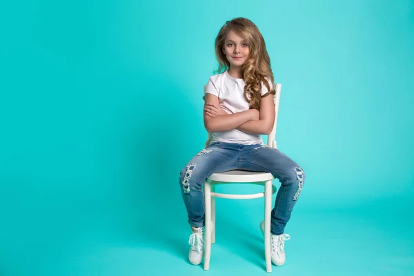 Piękne, wspaniałe trochę Blondynka w spodnie jeansowe siedzi na krześle na niebieskim tle — Zdjęcie stockowe