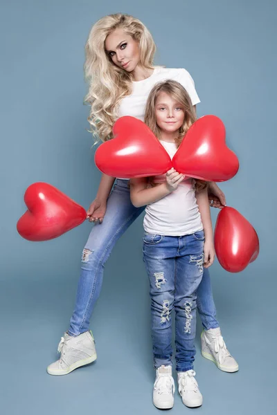 Vacker blond mor och dotter står på en blå bakgrund och innehav röda hjärtans ballonger — Stockfoto