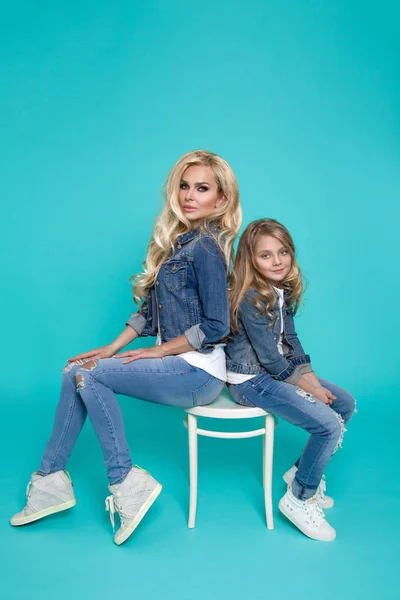 Fantastiska skönhet av en ung mor med en söt blond dotter som sitter på en stol på en blå bakgrund klädda i en denim jacka och byxor, kläder våren — Stockfoto