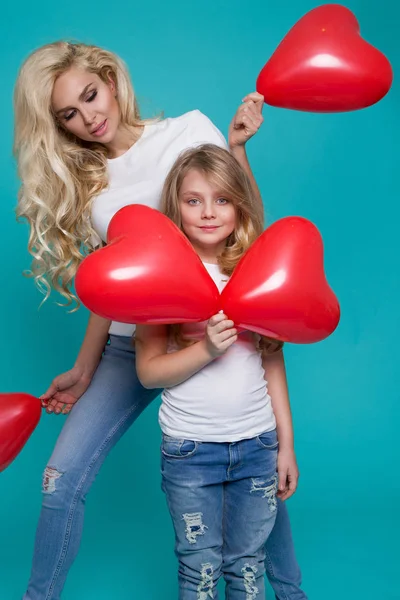 Schöne blonde Mutter und Tochter stehen auf blauem Hintergrund und halten rote Valentinusballons in der Hand — Stockfoto