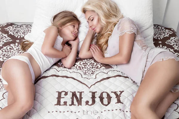 Porträtt av vacker blond kvinna mor och dotter på vackra ansikte och fantastiska ögon lögn sova på en säng i en elegant linne, krama varandra och ler glatt — Stockfoto