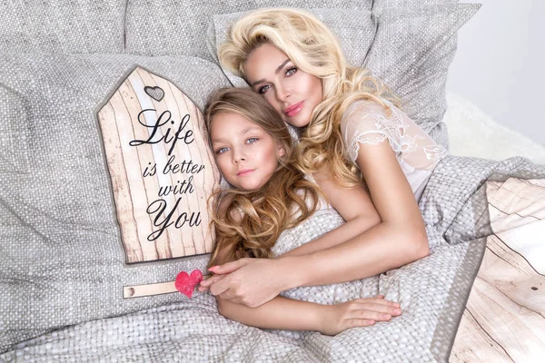 Portret van de mooie blonde vrouw moeder en dochter op de mooie gezicht en verbazingwekkend ogen liggen slapen op een bed in een elegante linnen, omhelzen elkaar en lachen vreugdevol — Stockfoto