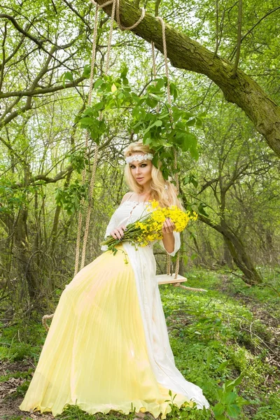 Εκπληκτική Ξανθιά σέξι γυναίκα φυσικό swing σε μια κούνια σε ένα δέντρο φορώντας ένα φόρεμα άνοιξη που κατέχουν ένα μπουκέτο της άνοιξης — Φωτογραφία Αρχείου
