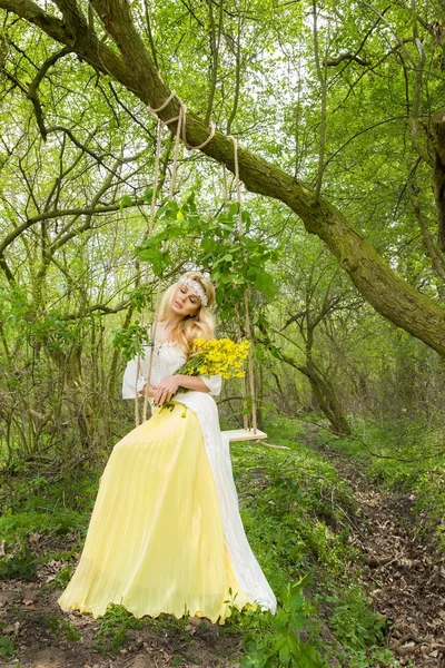 Splendida bionda sexy donna naturale altalena su un'altalena su un albero che indossa un abito primaverile che tiene un bouquet di primavera — Foto Stock
