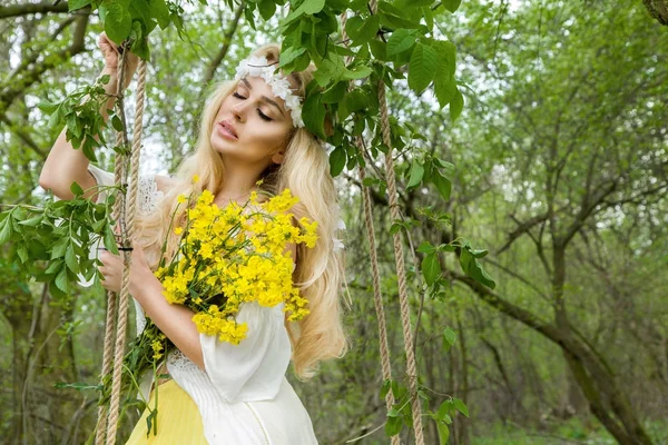 Mulher natural sexy loira deslumbrante balançar em um balanço em uma árvore usando um vestido de primavera segurando um buquê de primavera — Fotografia de Stock