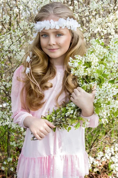 Schattig jong meisje met lang blond haar permanent in een weiland in krans van bloemen, holding van een boeket Lentebloemen en gekleed in lente kleding — Stockfoto