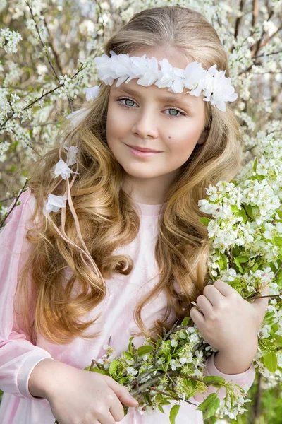 Schattig jong meisje met lang blond haar permanent in een weiland in krans van bloemen, holding van een boeket Lentebloemen en gekleed in lente kleding — Stockfoto