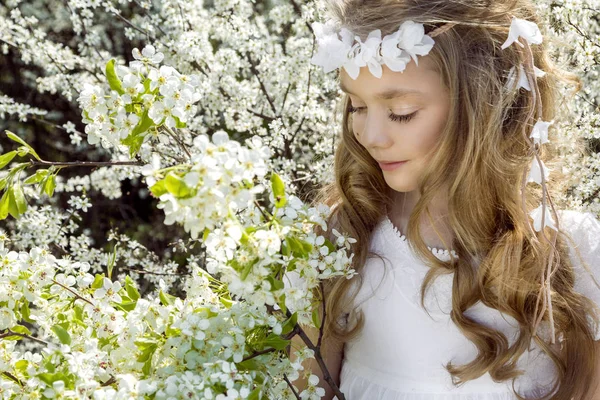 Menina bonito com longos cabelos loiros em pé em um prado em grinalda de flores, segurando um buquê de flores da primavera e vestido com roupas de primavera — Fotografia de Stock