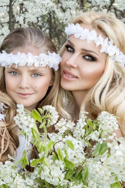 Beautiful young mother with her daughter dressed in spring clothes and wreaths of flowers stand on a meadow and snuggling — Stock Photo, Image