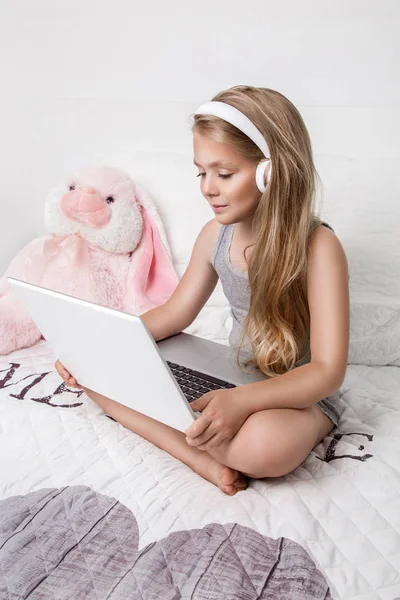 Gracious young beautiful blonde girl, little princess with long hair and blue eyes, the child lies in bed, listening to music, playing the game on a laptop computer and smiles joyfully — Stock Photo, Image