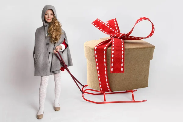 Menina bonita com longos cabelos loiros em pé sobre um fundo branco e segura um trenó com presentes — Fotografia de Stock