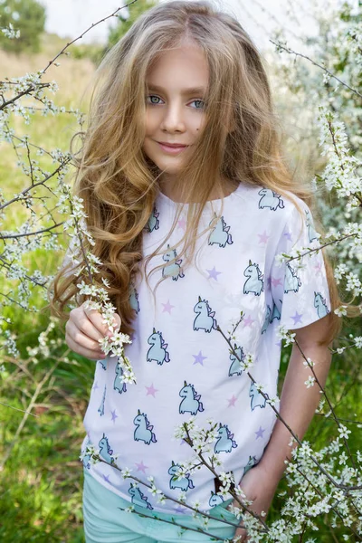 Menina bonito com longos cabelos loiros em pé em um prado em grinalda de flores, segurando um buquê de flores da primavera e vestido com roupas de primavera — Fotografia de Stock