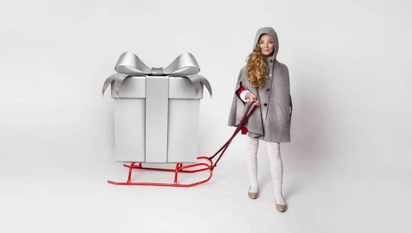 Pretty little girl with long blond hair standing on a white background and holds a sled with gifts — Stock Photo, Image