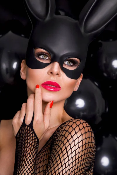 Mooie blonde sensuele vrouw met carnaval masker, staande op een achtergrond van zwarte ballonnen en sexy in de lens kijkt — Stockfoto