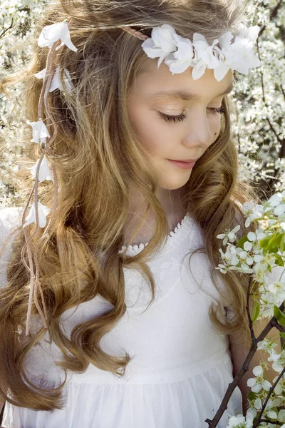 Menina bonito com longos cabelos loiros em pé em um prado em grinalda de flores, segurando um buquê de flores da primavera e vestido com roupas de primavera — Fotografia de Stock