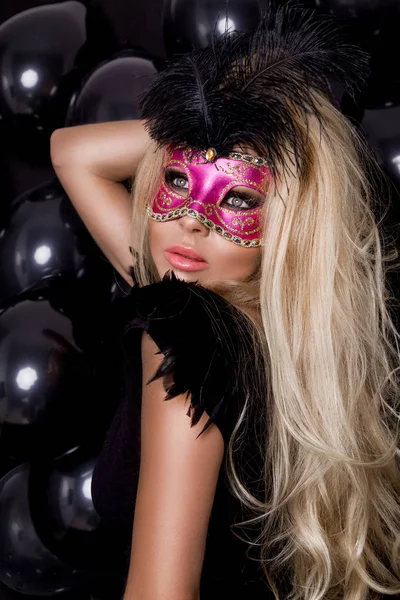 Schöne sinnliche blonde Frau mit Karnevalsmaske, stehend auf einem Hintergrund aus schwarzen Luftballons und sexy Blicken in die Linse — Stockfoto