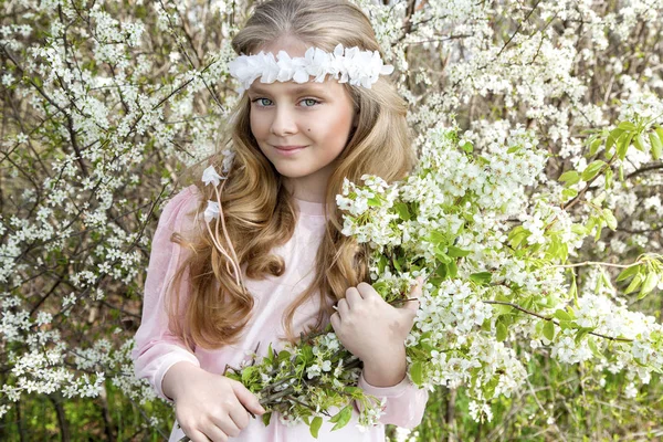 Χαριτωμένο νεαρή κοπέλα με μακριά ξανθά μαλλιά στέκεται σε ένα λιβάδι στο στεφάνι με λουλούδια, κρατώντας ένα μπουκέτο λουλούδια άνοιξη και ντυμένοι με ρούχα την άνοιξη — Φωτογραφία Αρχείου