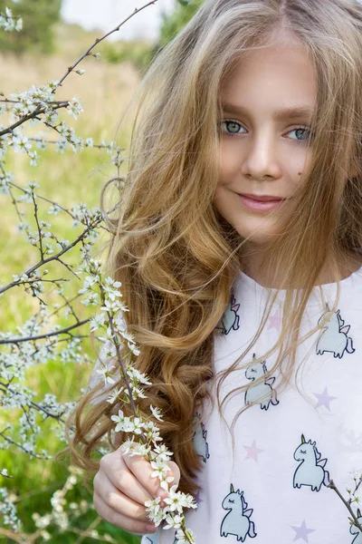 Menina bonito com longos cabelos loiros em pé em um prado em grinalda de flores, segurando um buquê de flores da primavera e vestido com roupas de primavera — Fotografia de Stock