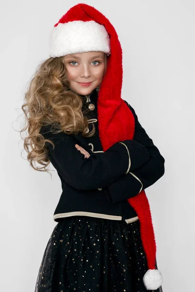 Stunning beautiful little girl with long blond, dressed in a red cap Santa Claus and elegant clothes stands on a white background and joyfully look into the lens — Stock Photo, Image