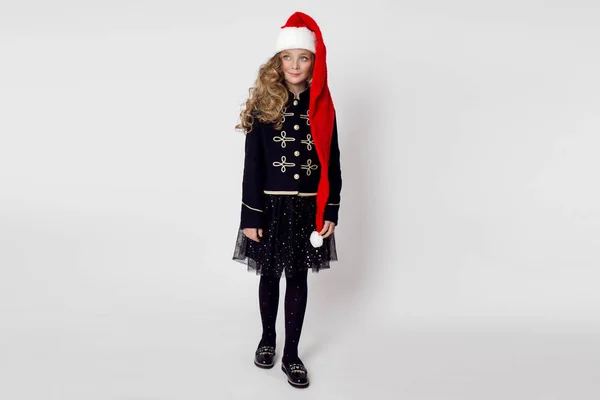 Потрясающая красивая маленькая девочка с длинной блондинкой, одетая в красную шапочку Санта-Клауса и элегантная одежда стоит на белом фоне и радостно смотрит в объектив — стоковое фото