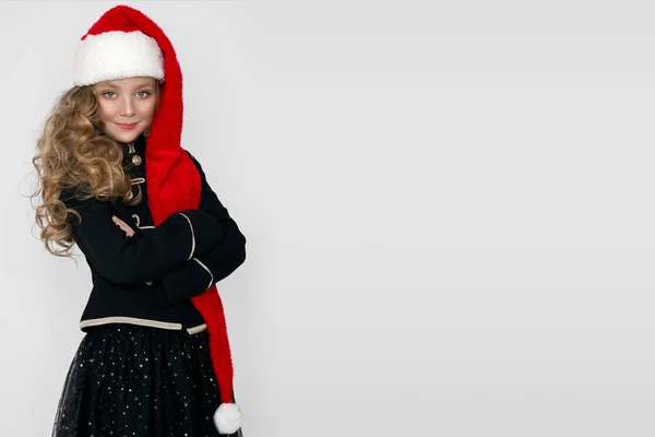 Menina bonita impressionante com loira longa, vestida com um chapéu vermelho Papai Noel e roupas elegantes fica em um fundo branco e olhar alegremente para a lente — Fotografia de Stock