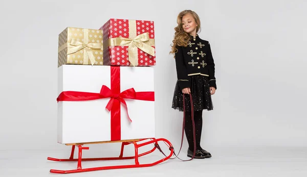 Hübsches kleines Mädchen mit langen blonden Haaren steht auf weißem Hintergrund und hält einen Schlitten mit Geschenken — Stockfoto