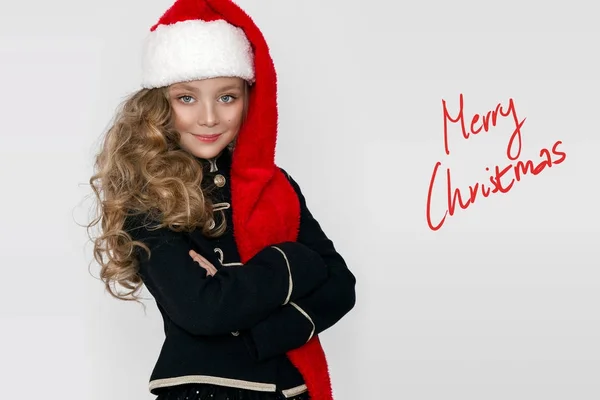 Atemberaubende schöne kleine Mädchen mit langen blonden, mit einer roten Mütze Weihnachtsmann und elegante Kleidung gekleidet steht auf einem weißen Hintergrund und schauen freudig in die Linse — Stockfoto