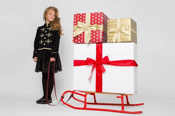 Досить маленька дівчинка з довгим світлим волоссям, що стоїть на білому тлі і тримає санки з подарунками — стокове фото