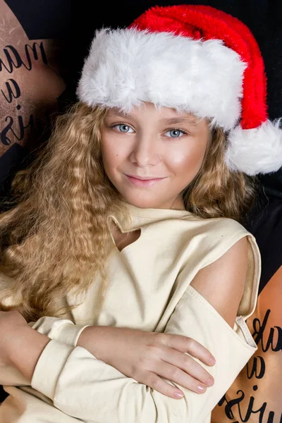 Krásná holčička má dlouhé blond vlasy, ležící na posteli v Santa Claus klobouk a radostně Podívejte se do objektivu v době Vánoc — Stock fotografie