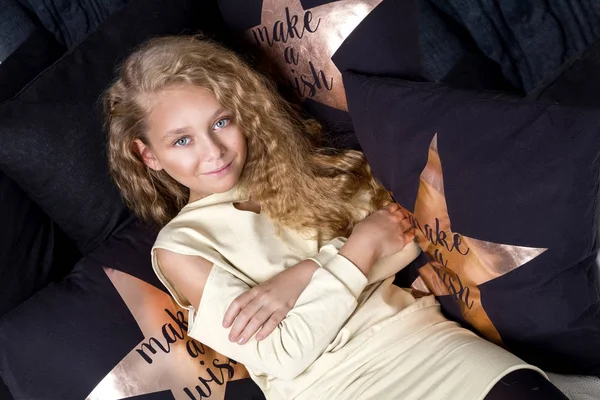 Gracious small a young girl with amazing eyes and blond hairs lies on the bed and in the background is black gold pillow with a star