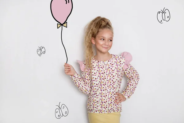 Εκπληκτικά όμορφο κοριτσάκι με μακριά ξανθά μαλλιά στέκεται πάνω σε ένα λευκό φόντο και κατέχει ένα μπαλόνι και τα λουλούδια στο χέρι του — Φωτογραφία Αρχείου