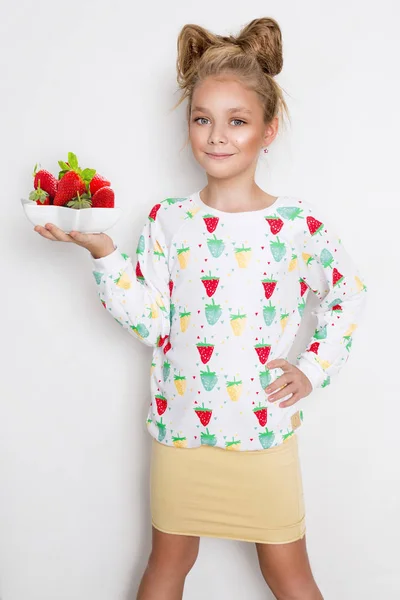 Niña graciosa con el pelo rubio y los ojos azules de pie sobre un fondo blanco con una sudadera y una fresa en la mano sosteniendo un tazón con fresas frescas — Foto de Stock