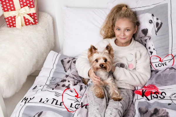 Потрясающая красивая маленькая девочка с длинными светлыми волосами лежит на постельном белье в печатных собаках и лежит рядом со своей собакой Йоркширский терьер радостно смотрит в объектив — стоковое фото