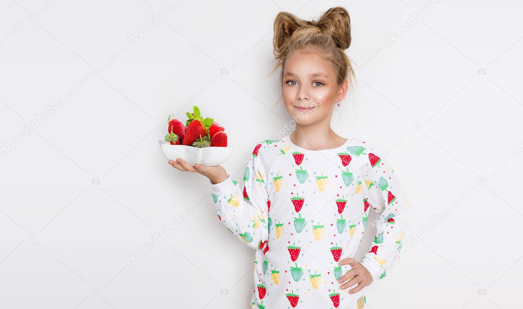 Gracious little girl with blond hair and blue eyes standing on a white background wearing a sweatshirt and a strawberry in his hand holding a bowl with fresh strawberries