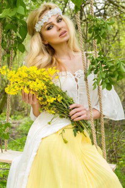 Bir buket bahar tutan bir Bahar elbise giyen bir ağaç salıncakta çarpıcı sarışın seksi doğal kadın salıncakta