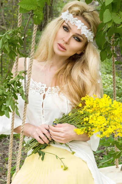 Splendida bionda sexy donna naturale altalena su un'altalena su un albero che indossa un abito primaverile che tiene un bouquet di primavera — Foto Stock