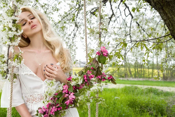 Fantastisk Blond sexig naturlig Kvinna gunga på en gunga på ett träd som bär en klänning för våren som håller en bukett av våren — Stockfoto