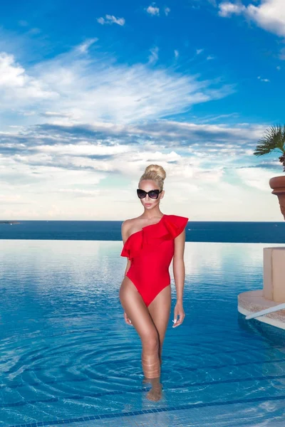 Krásná blondýnka model s úžasné tělo stojící v bazénu v elegantní červené plavky a v pozadí je úžasný výhled na moře a obloha. — Stock fotografie