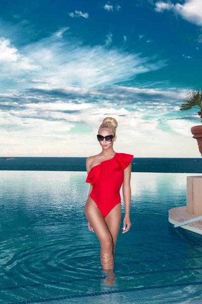 Stunning blonde female model model with amazing body standing in the pool in an elegant red swimsuit and in the background is amazing view of the sea and sky. — Stock Photo, Image