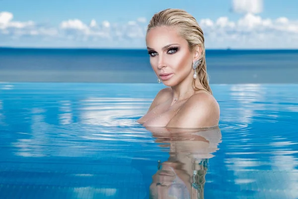 Islak saç ve zarif makyaj takı, kristal ve ıslak çıplak vücut ile küpeler harika manzaralı lüks otel, bir havuzda oturan güzel sarışın kadın model