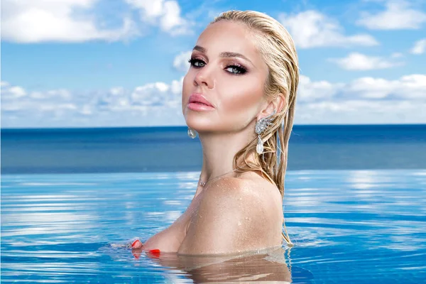 Krásná blondýnka model s mokrými vlasy a elegantní make-up sedí v bazénu s nádherným výhledem v luxusním hotelu, nosí šperky, náušnice s krystaly a mokré nahé tělo — Stock fotografie