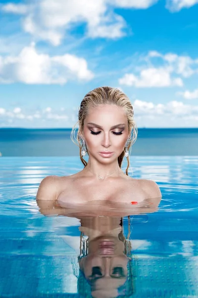 Islak saç ve zarif makyaj takı, kristal ve ıslak çıplak vücut ile küpeler harika manzaralı lüks otel, bir havuzda oturan güzel sarışın kadın model