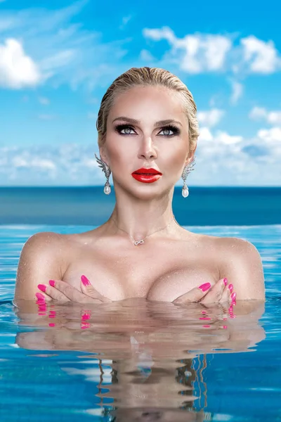 Piękna Kobieta blonde model z mokrych włosów i elegancki makijaż siedzi w basenie ze wspaniałym widokiem na luksusowy hotel, noszenia biżuterii, kolczyki z kryształów i mokre nagie ciało — Zdjęcie stockowe