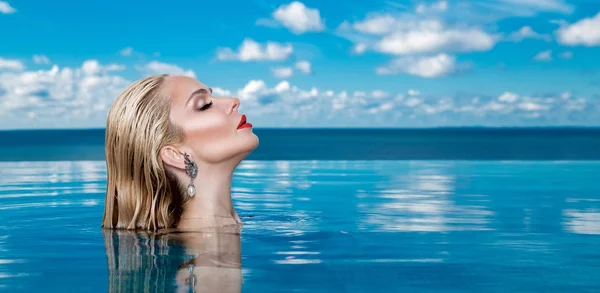 Beautiful blonde woman model with wet hair and elegant makeup sitting in a pool with amazing views in a luxury hotel, wearing jewelry, earrings with the crystals and wet naked body — Stock Photo, Image