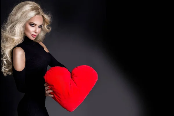 Krásná ohromující žena s dlouhé blond vlasy stojící na černém pozadí oblečený v černých šatech a drží v rukou červené srdce na Valentýna — Stock fotografie