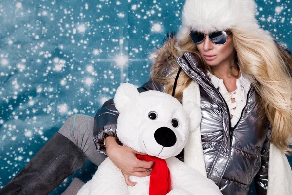 Belle femme magnifique avec de longs cheveux blonds et un visage parfait habillé en vêtements d'hiver, veste chaude argentée et casquette en fourrure et lunettes de soleil argentées sur fond d'hiver — Photo