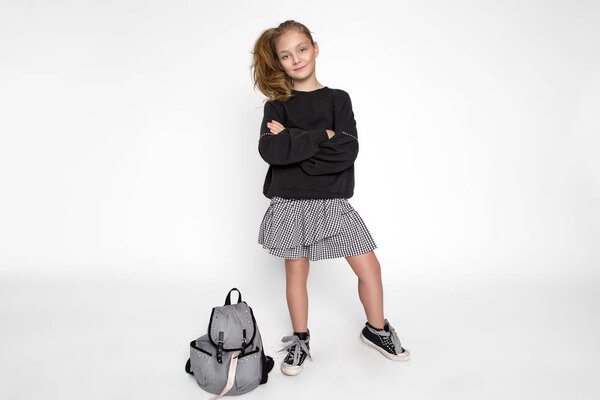 Cute little girl dressed in school clothes and school backpack sitting on white background